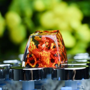 May include: A custom keycap featuring a small orange Charmander figure surrounded by a fiery, lava-like design. The keycap is clear, allowing the design to be visible. It sits atop a keyboard with silver keycaps.