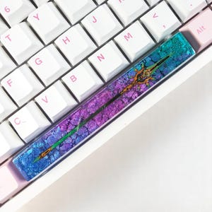 Pode incluir: Um teclado com uma tecla personalizada. A tecla é feita de resina e tem um esquema de cores azul, roxo e verde. Apresenta um design dourado de uma lança.