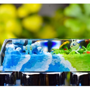 Peut inclure: Une touche de clavier en résine transparente représentant une scène océanique miniature avec des vagues bleues et une petite créature bleue. Le côté droit montre une zone herbeuse verte avec une créature blanche et verte et des détails rouges.