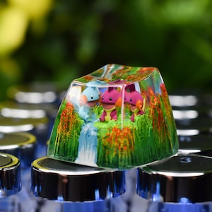 May include: A custom keycap with a colourful scene depicting three cartoon characters, a waterfall, and vibrant foliage. The clear keycap allows the design to be visible. The keycap is set on a keyboard.