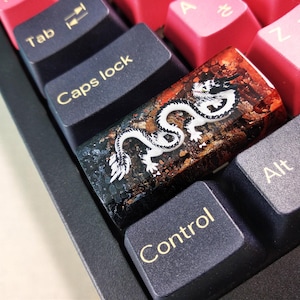 Puede incluir: Un tapón de tecla de dragón blanco y negro con una base de resina marrón, naranja y dorada. El tapón de tecla está en un teclado negro con las teclas "Bloq Mayús", "Control" y "Alt" visibles.