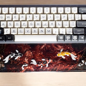 May include: A black and white keyboard with a white keycap set and a black and red koi fish themed wrist rest. The wrist rest has five koi fish in various colors, including black, white, orange, and yellow.