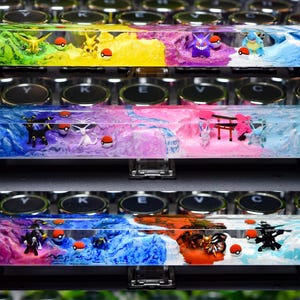 Könnte beinhalten: Drei Harz-Tastenkappen mit verschiedenen Pokémon-Charakteren und Landschaften. Die erste Taste ist grün, gelb und blau mit einem Pikachu, einem Relaxo und einem Blastoise. Die zweite Taste ist rosa, lila und blau mit einem Evoli, einem Gengar und einem Aquana. Die dritte Taste ist rot, orange und blau mit einem Glumanda, einem Flamara und einem Blitza.