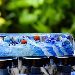 Peut inclure: Une touche de clavier personnalisée avec un diorama miniature. La touche transparente présente une scène océanique bleue et blanche, avec de petites figurines de personnages Pokémon et des Poké Balls. La touche est montée sur un clavier mécanique.