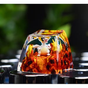 May include: A custom keycap featuring a white and orange dragon figure with green wings, set against a fiery background. The keycap is encased in clear resin and sits atop a black keyboard.