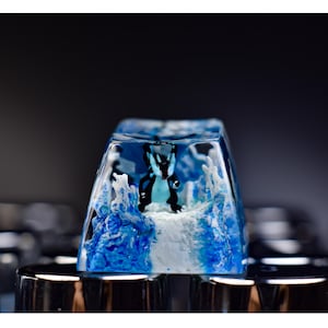 May include: A custom keycap with a blue and white design and a black figure. The keycap is made of clear resin, with a detailed, artistic design. The keycap is part of a keyboard.