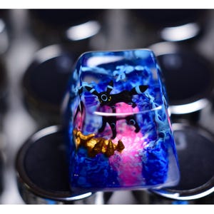 Könnte beinhalten: Ein blauer und rosa Harz-Tastenkappen mit einem schwarzen und weißen Wesen darin. Die Tastenkappe ist von anderen Tastenkappen umgeben.