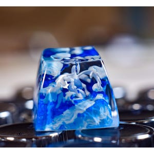 May include: A blue and white resin keycap with a swirling cloud design. The keycap is made of clear resin with white and blue swirls inside.