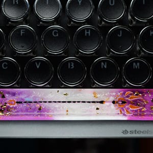 Pode incluir: Teclado com teclas redondas pretas. Barra de espaço personalizada com um design roxo, branco e dourado. A barra de espaço tem um design intrincado com flocos de ouro e um detalhe laranja. Teclado SteelSeries.