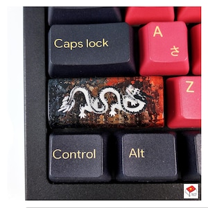 Peut inclure: Un capuchon de touche de clavier représentant un dragon noir et blanc sur une base en résine rouge, orange et marron. Le capuchon de touche est sur un clavier noir avec des lettres dorées.
