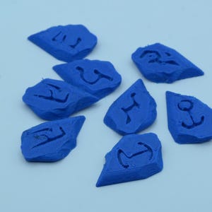 Puede incluir: Ocho piedras azules de forma irregular, cada una con un s&iacute;mbolo diferente tallado en su superficie. Los s&iacute;mbolos incluyen un ancla, una letra estilizada y otros dise&ntilde;os abstractos. Las piedras est&aacute;n dispuestas sobre una superficie blanca.