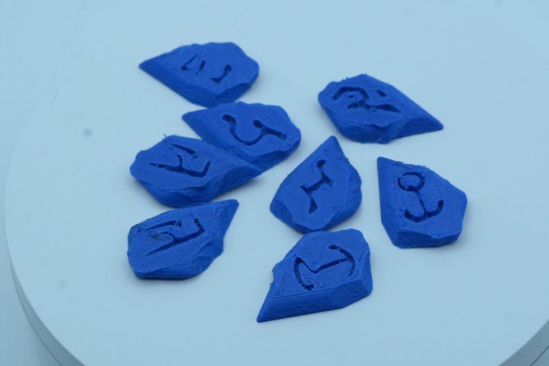 Puede incluir: Ocho piedras azules de forma irregular, cada una con un s&iacute;mbolo diferente tallado en su superficie. Los s&iacute;mbolos incluyen un ancla, una letra estilizada y otros dise&ntilde;os abstractos. Las piedras est&aacute;n dispuestas sobre una superficie blanca.