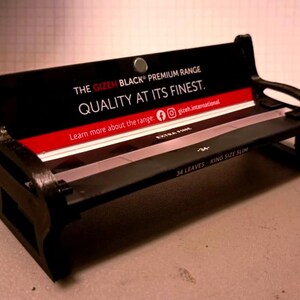 Può includere: Una piccola panchina nera con una striscia rossa e bianca, con il testo "THE GIZEH BLACK PREMIUM RANGE QUALITY AT ITS FINEST." La panchina presenta anche la scritta "EXTRA FINE" e "34 LEAVES KING SIZE SLIM."