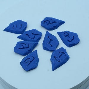 Puede incluir: Una colecci&oacute;n de ocho piedras azules de forma irregular, cada una con un s&iacute;mbolo diferente tallado en su superficie. Las piedras est&aacute;n dispuestas sobre una superficie circular blanca. Los s&iacute;mbolos parecen n&aacute;uticos o antiguos.