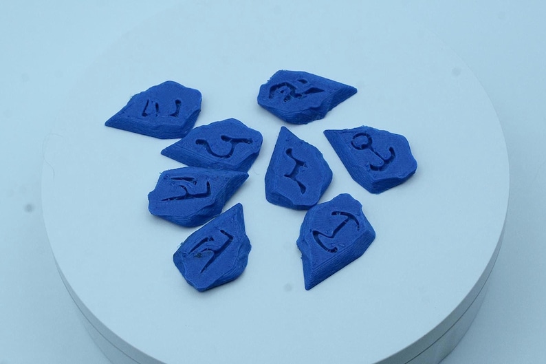 Puede incluir: Una colecci&oacute;n de ocho piedras azules de forma irregular, cada una con un s&iacute;mbolo diferente tallado en su superficie. Las piedras est&aacute;n dispuestas sobre una superficie circular blanca. Los s&iacute;mbolos parecen n&aacute;uticos o antiguos.