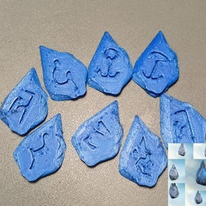 Puede incluir: Nueve piedras azules de forma irregular con s&iacute;mbolos grabados. Las piedras est&aacute;n dispuestas sobre una superficie neutra. Una imagen m&aacute;s peque&ntilde;a de piedras similares est&aacute; en la esquina inferior derecha. Los s&iacute;mbolos parecen ser de tem&aacute;tica n&aacute;utica o mar&iacute;tima.