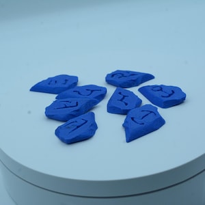 Puede incluir: Un conjunto de ocho piedras azules de forma irregular, cada una con un s&iacute;mbolo tallado. Las piedras est&aacute;n dispuestas sobre una superficie circular blanca. Los s&iacute;mbolos parecen runas u otras marcas antiguas.