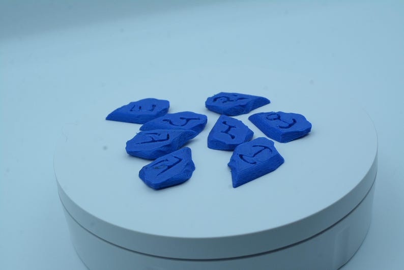 Puede incluir: Un conjunto de ocho piedras azules de forma irregular, cada una con un s&iacute;mbolo tallado. Las piedras est&aacute;n dispuestas sobre una superficie circular blanca. Los s&iacute;mbolos parecen runas u otras marcas antiguas.