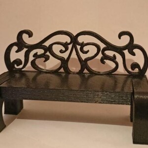 May include: A small black bench with an elaborate, scrolled back. The bench has a rectangular seat and curved legs. The back features a symmetrical design of swirling patterns. Suitable for dollhouses or miniature displays, this bench is a decorative item.