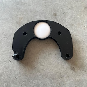 May include: A black, curved object with a circular white center and two small holes. The object has a smooth, matte finish and appears to be made of plastic or a similar material. The design is symmetrical, with a rounded shape.