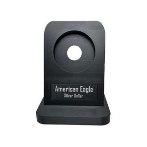 Puede incluir: Soporte de exhibición de monedas negro con un recorte circular para una moneda. El soporte tiene una etiqueta que dice "American Eagle Silver Dollar".