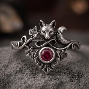 May include: A silver ring featuring a sculpted fox head, a crescent moon, and ivy leaf motifs. The centre of the ring is set with a round, deep red gemstone. The metal has an oxidised finish that highlights the intricate details of the fox and foliage.