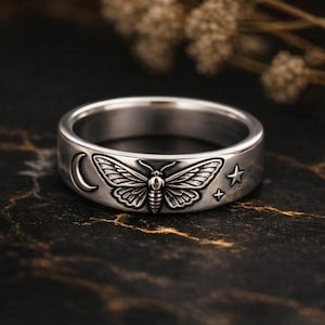 May include: Silver ring with a detailed moth, crescent moon, and star design. The ring has a wide band and a vintage, antique style. The moth is the central focus, with intricate wing and body details. The ring is likely made of sterling silver.