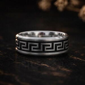 May include: A silver ring featuring a Greek key design around the band. The ring has a flat top and a slightly rounded edge. The geometric pattern is in a darker shade of silver, creating a contrast.