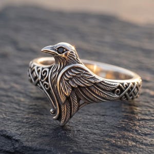 May include: A silver-toned ring featuring a sculpted raven design with a small black stone set as the eye. The band of the ring is decorated with intricate, repeating geometric patterns that extend along the sides of the bird figure.