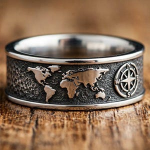 May include: A silver-toned metal ring featuring a textured, dark grey band engraved with a world map and a compass rose. The continents are raised and polished with a metallic finish, contrasting against the recessed, dark, hammered-style background.