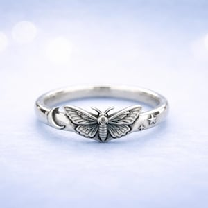 May include: A silver ring featuring a detailed moth design at the center. The ring has a crescent moon and stars on either side of the moth. The ring is set against a soft, blurred background.