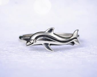 May include: A silver ring featuring a detailed dolphin design. The dolphin is sculpted with visible fins and a smiling face, with a small accent on the eye. The ring band is smooth and simple, creating a nautical theme.