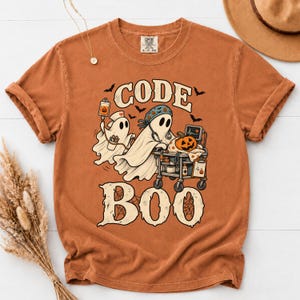 May include: A burnt orange t-shirt featuring a graphic of two ghosts dressed as medical professionals with a medical cart, a jack-o'-lantern, and bats. The text reads CODE BOO in large, distressed, cream-colored lettering above and below the ghosts.