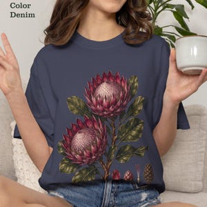 Könnte beinhalten: Ein denimfarbenes T-Shirt mit einer botanischen Illustration von zwei großen, dunkelrosa Protea-Blüten mit dunkelgrünen Blättern und Stielen. Die Grafik enthält Details von Knospen und Samenkapseln auf einem einfarbigen, dunkelblauen Hintergrund.