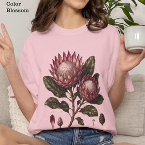 Può includere: Una t-shirt rosa chiaro con un'illustrazione botanica di due grandi fiori di protea con foglie verde scuro e piccoli elementi floreali nella parte inferiore. Il testo Color Blossom è stampato nell'angolo in alto a sinistra dell'immagine.