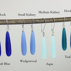 May include: Six pairs of teardrop-shaped glass earrings in various shades of blue, including cobalt blue, wedgewood, aqua, and teal. The earrings are displayed on a wooden dowel with the hook style and kidney wire style earring findings.