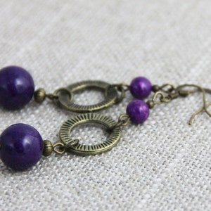 May include: A pair of drop earrings featuring large, smooth, deep purple spherical beads paired with smaller matching purple beads. The beads are connected by antique bronze-toned metal rings with a textured, radial line pattern and hooks.