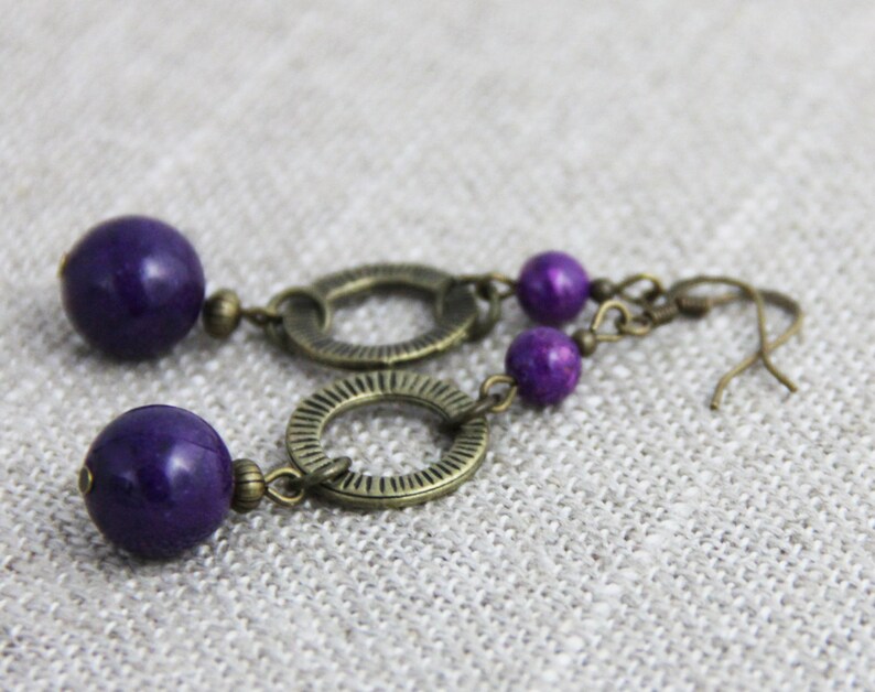 May include: A pair of drop earrings featuring large, smooth, deep purple spherical beads paired with smaller matching purple beads. The beads are connected by antique bronze-toned metal rings with a textured, radial line pattern and hooks.