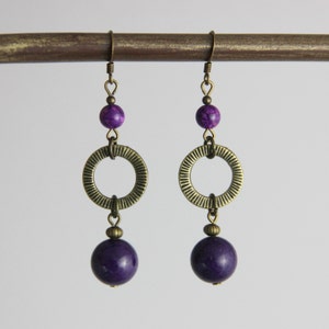 May include: A pair of drop earrings featuring antique brass hooks, a small purple marbled bead, a textured brass ring with radial lines, and a larger smooth dark purple spherical bead at the base, all connected by brass jump rings and findings.
