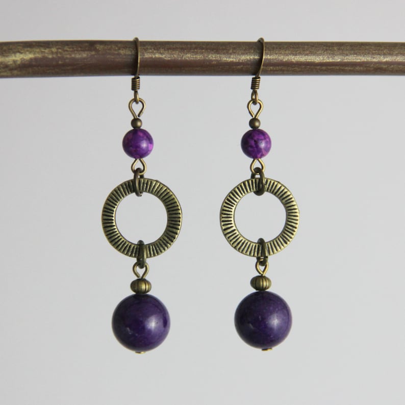 May include: A pair of drop earrings featuring antique brass hooks, a small purple marbled bead, a textured brass ring with radial lines, and a larger smooth dark purple spherical bead at the base, all connected by brass jump rings and findings.