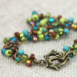 May include: A beaded bracelet with a mix of green, brown, blue, and iridescent beads. The bracelet has a decorative bronze clasp.