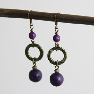 May include: A pair of dangle earrings featuring antique brass hooks, a small purple bead, a textured brass ring, and a larger round purple bead at the bottom. The earrings are suspended from a dark brown horizontal rod against a plain white background.