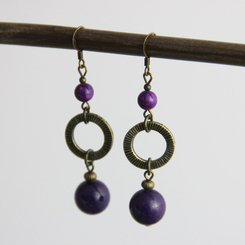 May include: A pair of dangle earrings featuring antique brass hooks, a small purple bead, a textured brass ring, and a larger round purple bead at the bottom. The earrings are suspended from a dark brown horizontal rod against a plain white background.