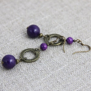 May include: A pair of drop earrings featuring deep purple spherical beads and antique bronze-toned metal components. Each earring consists of a large purple bead, a textured circular metal ring, a smaller purple bead, and a hook-style ear wire.