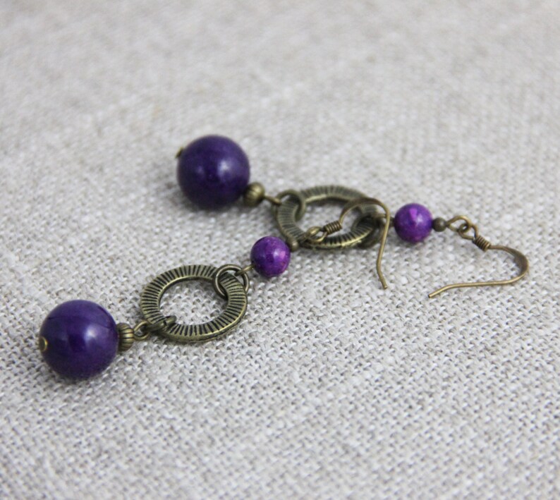 May include: A pair of drop earrings featuring deep purple spherical beads and antique bronze-toned metal components. Each earring consists of a large purple bead, a textured circular metal ring, a smaller purple bead, and a hook-style ear wire.