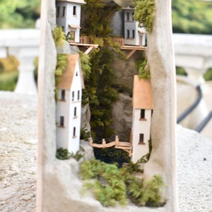 Könnte beinhalten: Eine Miniatur-Dorfszene mit weißen Häusern und einer kleinen Brücke über einem Teich, alles in einen Holzrahmen eingebaut.