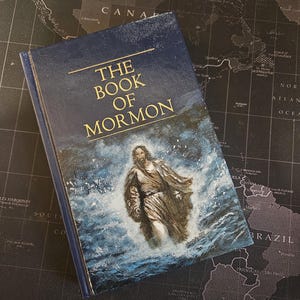 May include: A dark blue hardback book titled The Book of Mormon in gold lettering. The cover features a painted illustration of a figure walking through turbulent, splashing blue and white water, with a textured finish across the entire surface.