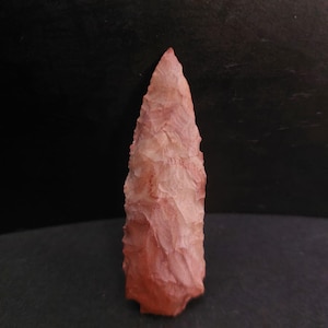 Peut inclure: Une pointe de flèche rose rougeâtre, taillée à la main, avec une pointe acérée et une surface texturée. L'ancien artefact est présenté sur un fond sombre, mettant en valeur son savoir-faire et ses variations de couleurs.