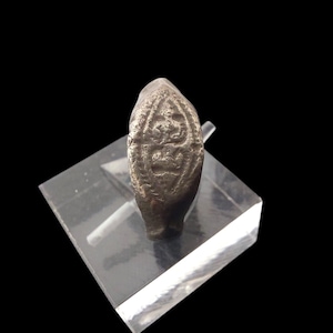 Puede incluir: Un anillo plateado con una cara ovalada con un diseño en relieve. El anillo se exhibe sobre un soporte transparente e inclinado sobre un fondo negro. El diseño en relieve parece ser una figura o símbolo estilizado.