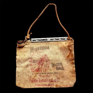 May include: A vintage canvas water bag with a rope handle and metal top closure. The bag features a faded red graphic of a person wearing a headdress, the text Minnequa Water Bag, and Pueblo Tent & Awning Co, Pueblo, Colorado printed on it.